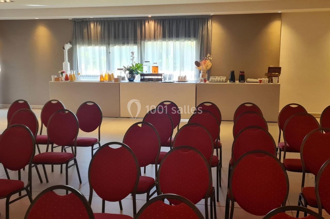 Salle de conférence avec des rangées de chaises rouges et une table au fond proposant des boissons et collations.