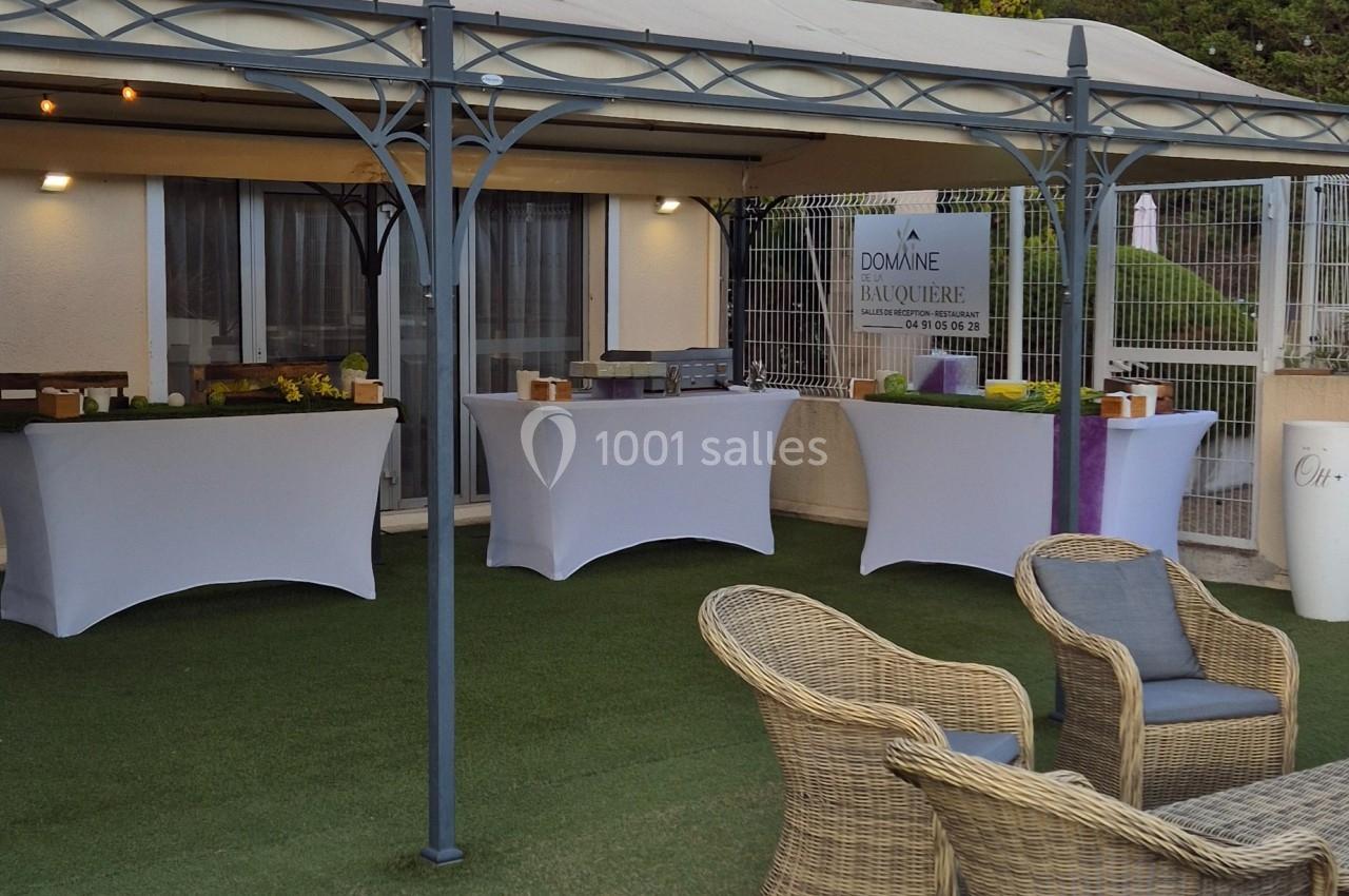 Tables habillées de nappes blanches sous une pergola, avec chaises en rotin au premier plan, dans un espace extérieur.