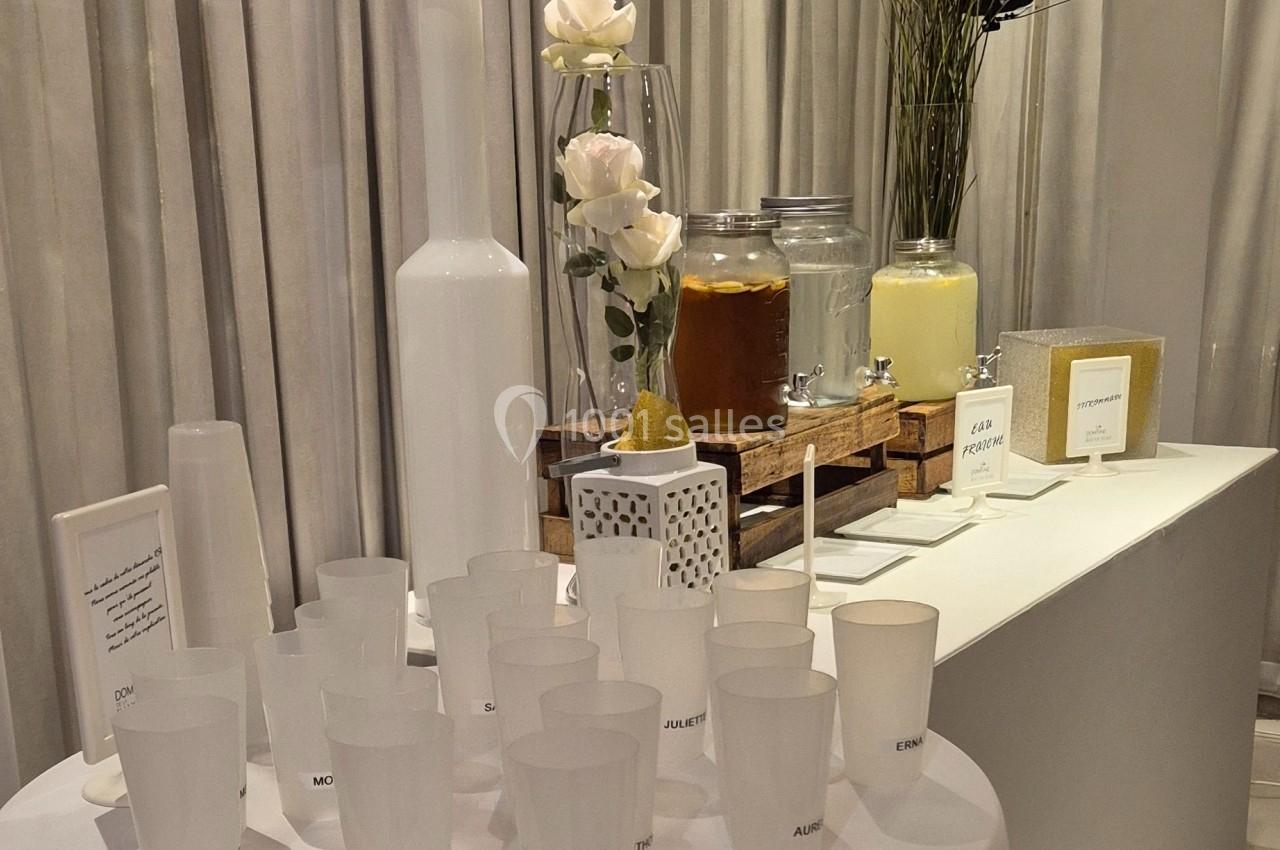 Table avec des boissons en distributeurs, verres en plastique translucides et décorations florales blanches.