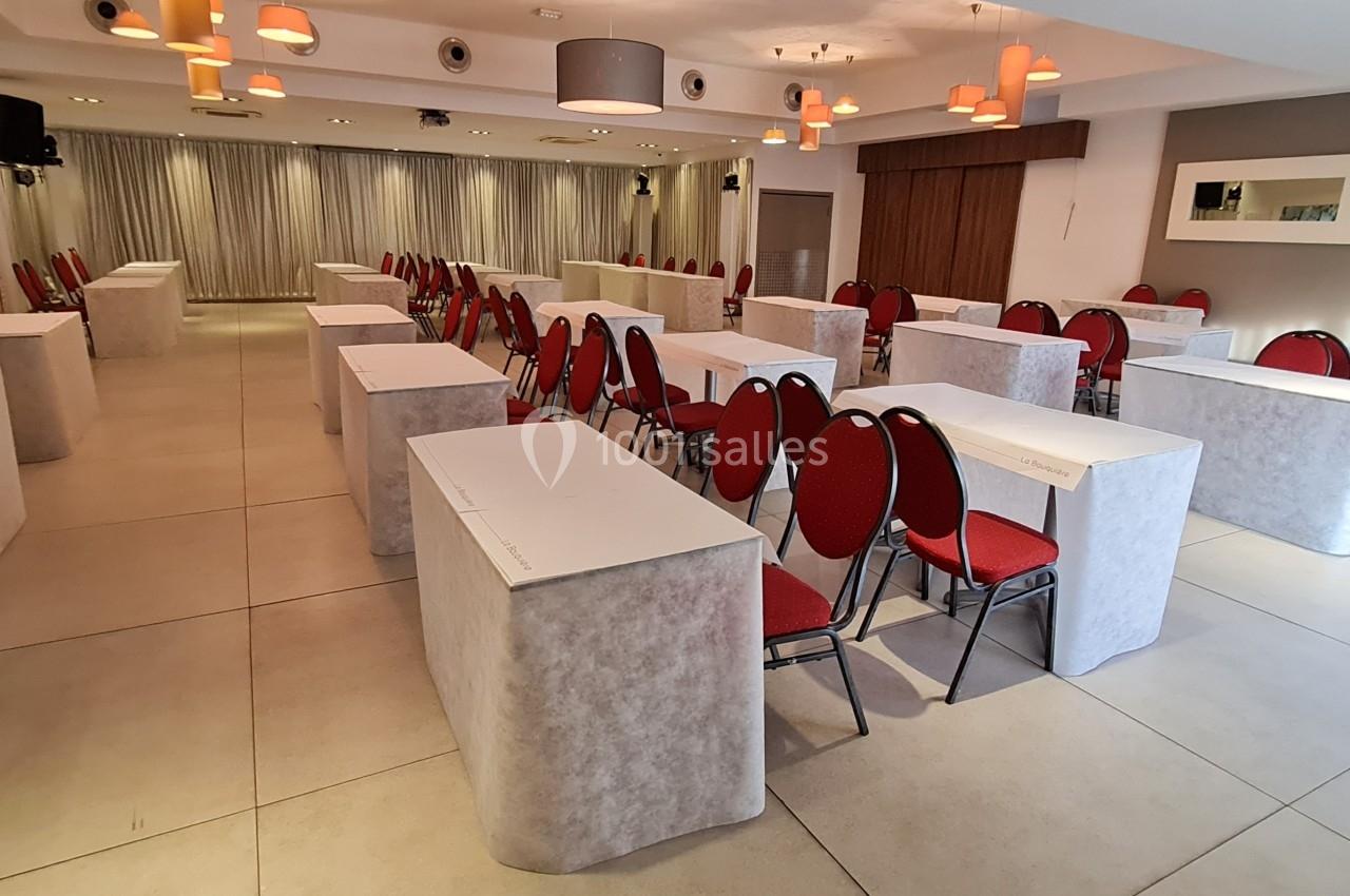 Salle de conférence lumineuse avec tables rectangulaires couvertes de nappes blanches et chaises rouges alignées.