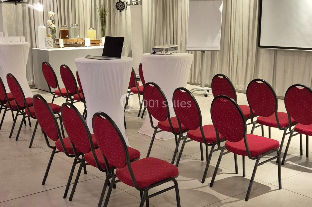 Salle de conférence avec des chaises rouges alignées, des tables hautes blanches et un projecteur installé.
