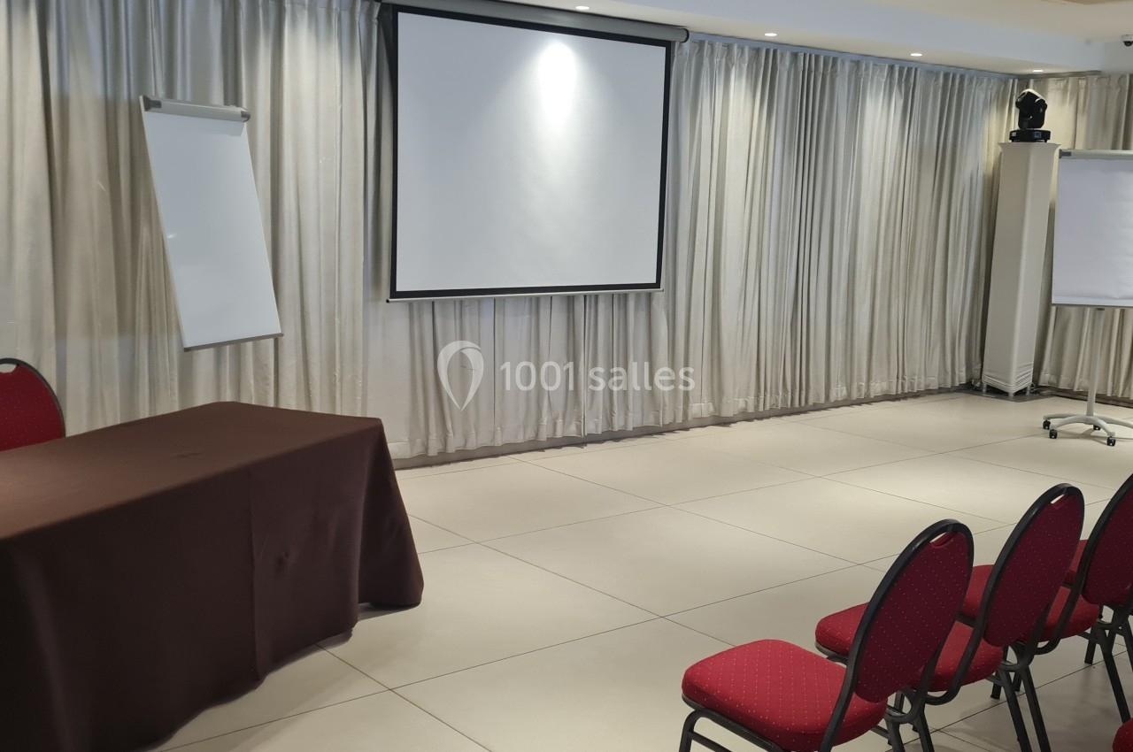 Salle de conférence avec écran de projection, deux chevalets de papier, table recouverte d'une nappe et rangée de chaises…