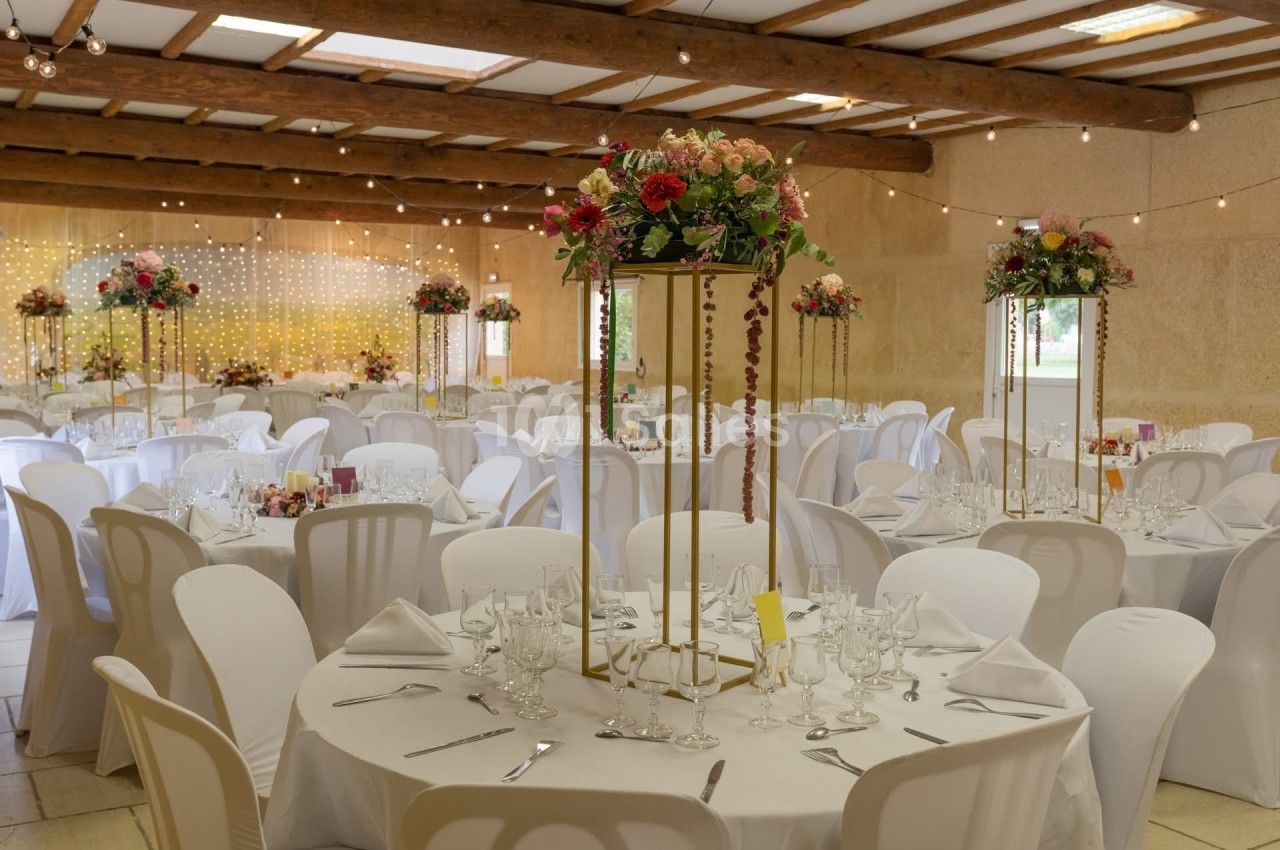 Salle de réception décorée avec des tables rondes, nappes blanches, centres de table fleuris et guirlandes lumineuses.
