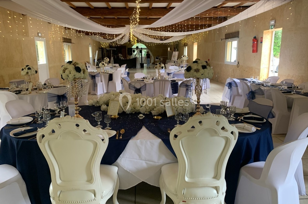 Salle de réception décorée avec des tables rondes élégantes, nappes bleues, chaises blanches et guirlandes lumineuses…