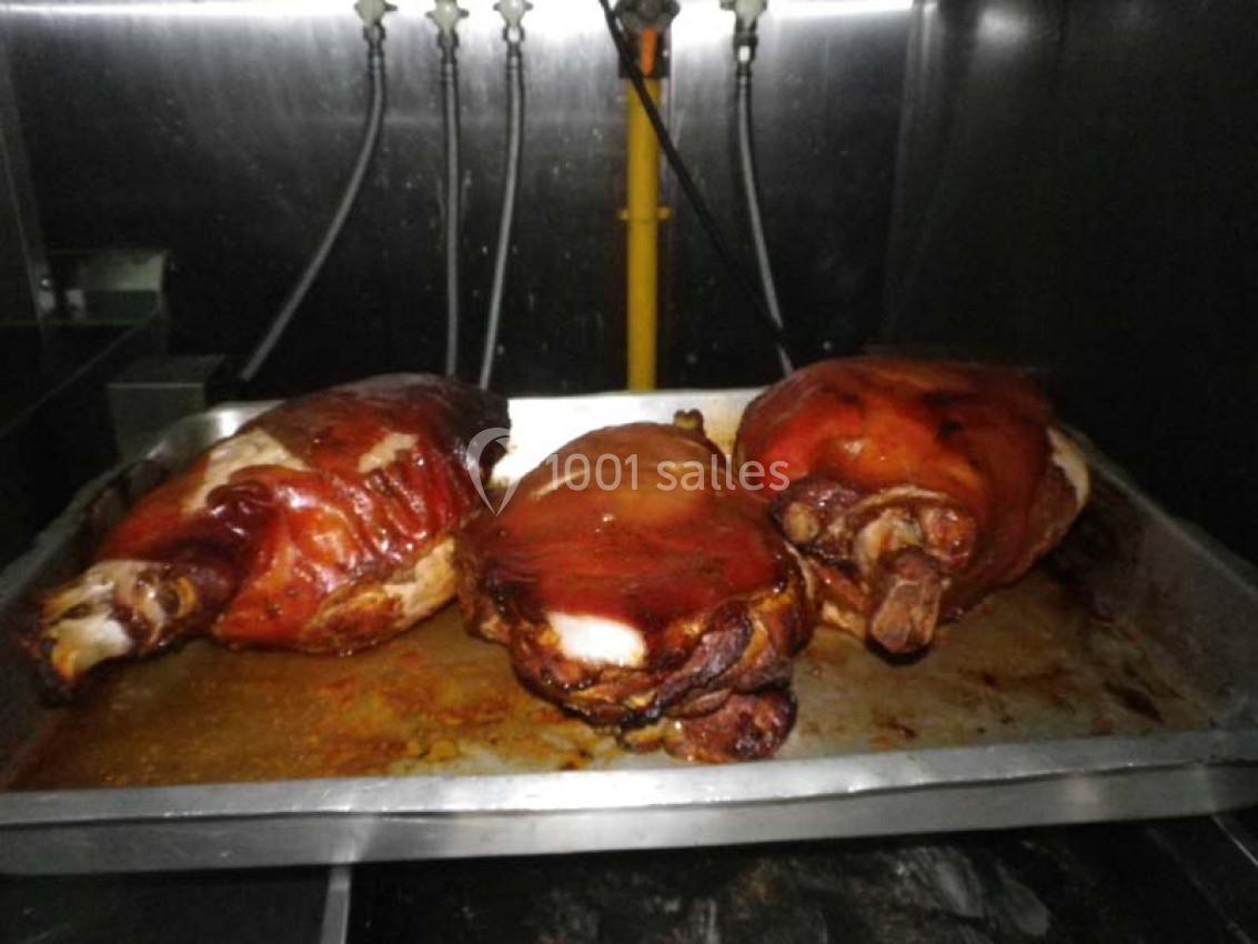 Trois jambons rôtis avec une peau dorée reposent dans un plat métallique dans une cuisine industrielle.