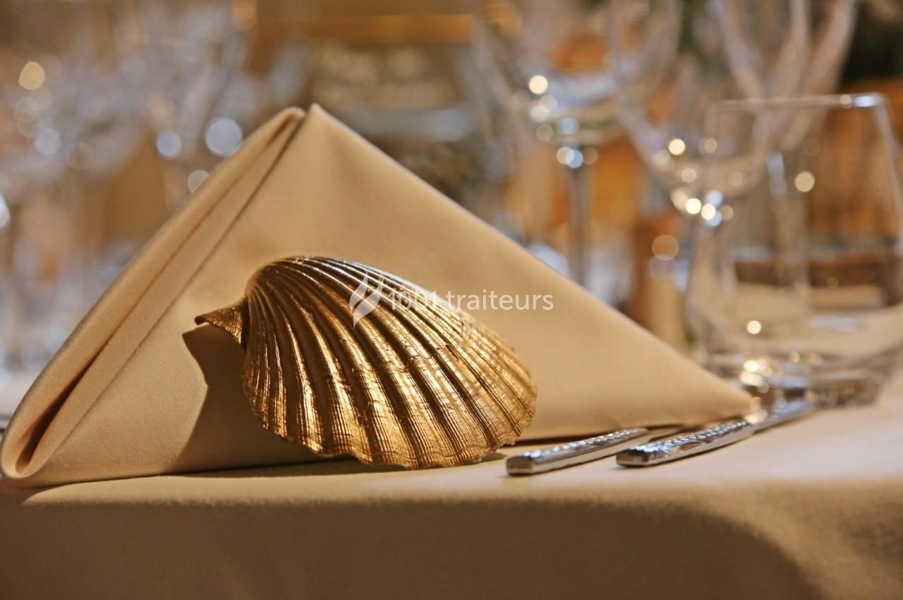 Serviette pliée en triangle décorée d'une coquille dorée sur une table dressée avec des verres et des couverts.