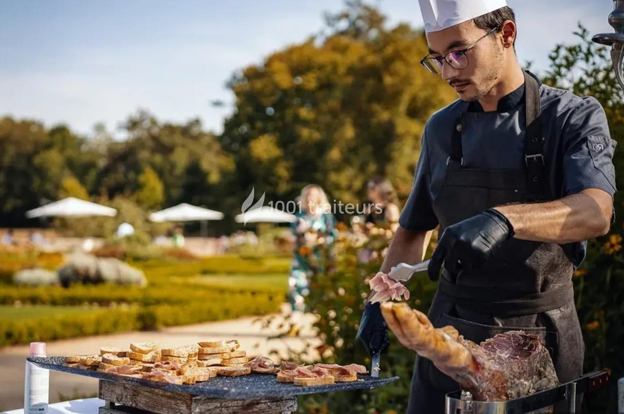 Un chef prépare des tranches de jambon et de pain grillé en extérieur, dans un jardin par une journée ensoleillée.