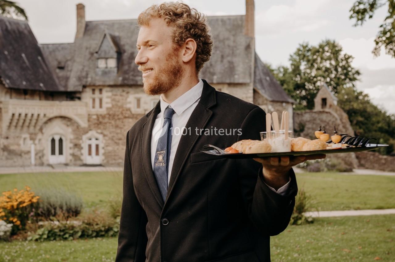 Serveur en costume portant un plateau de canapés dans un jardin devant un bâtiment en pierre de style ancien.