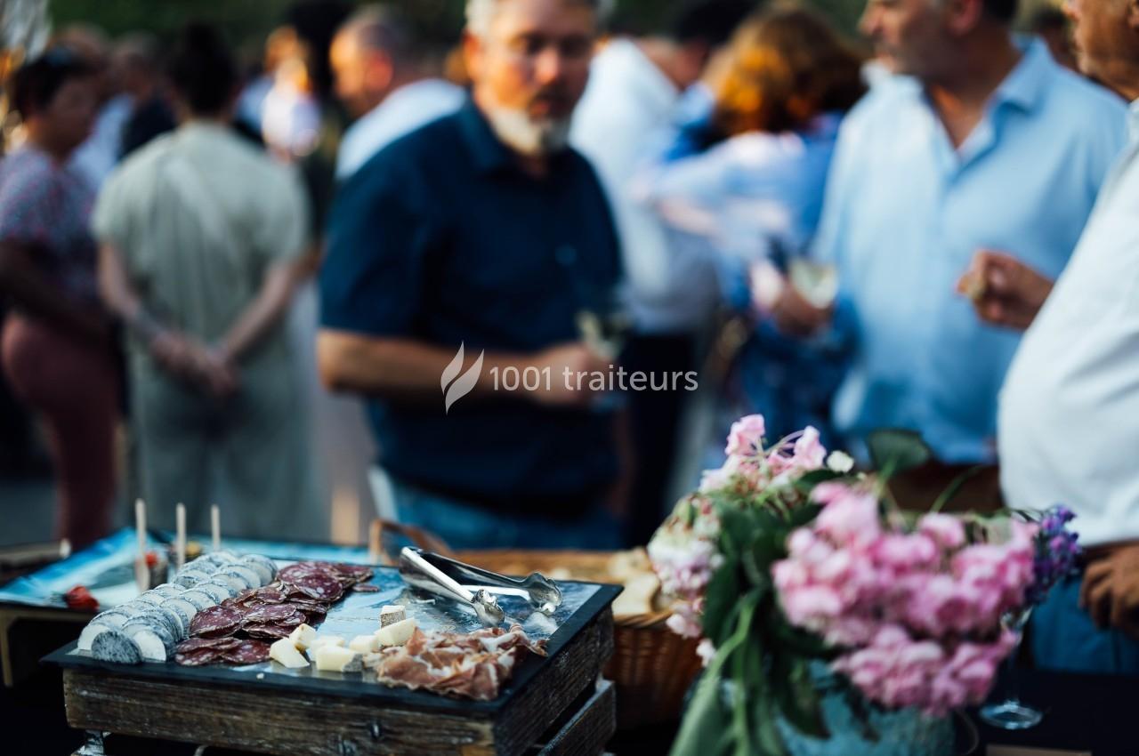 Plateau de charcuterie et fromages avec des fleurs roses au premier plan, des personnes floues en arrière-plan.