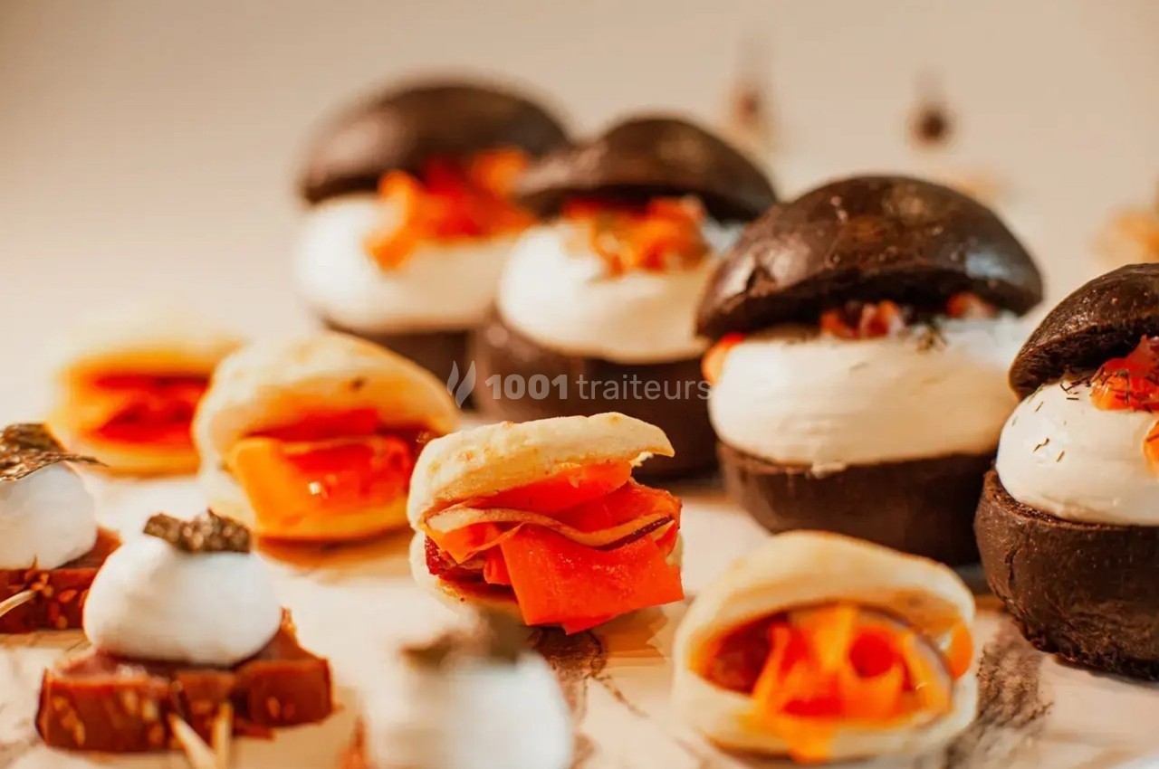 Assortiment de mini-burgers noirs et bouchées garnies de légumes et fromage, disposés sur une table.