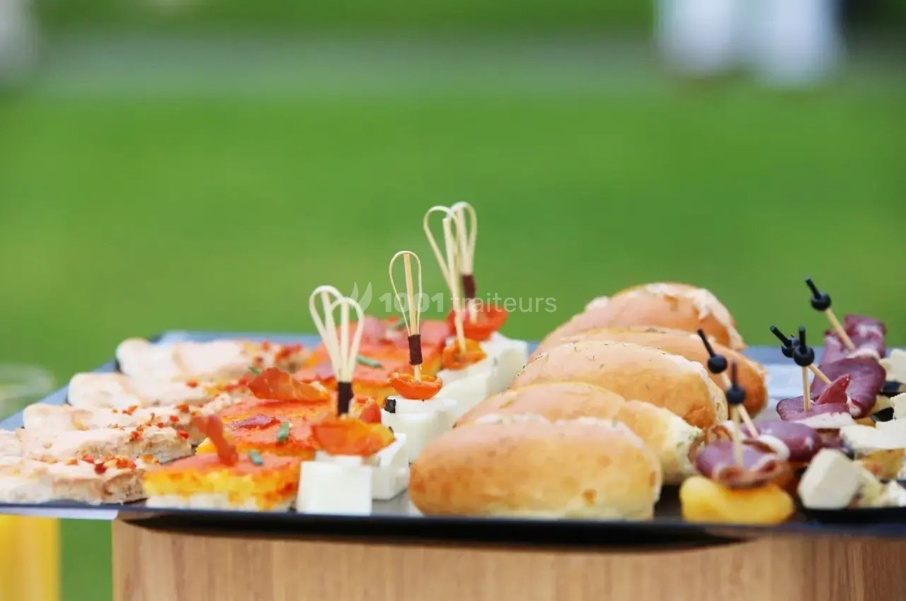 Plateau de bouchées apéritives variées comprenant mini-sandwichs, brochettes et toasts, présenté en extérieur.