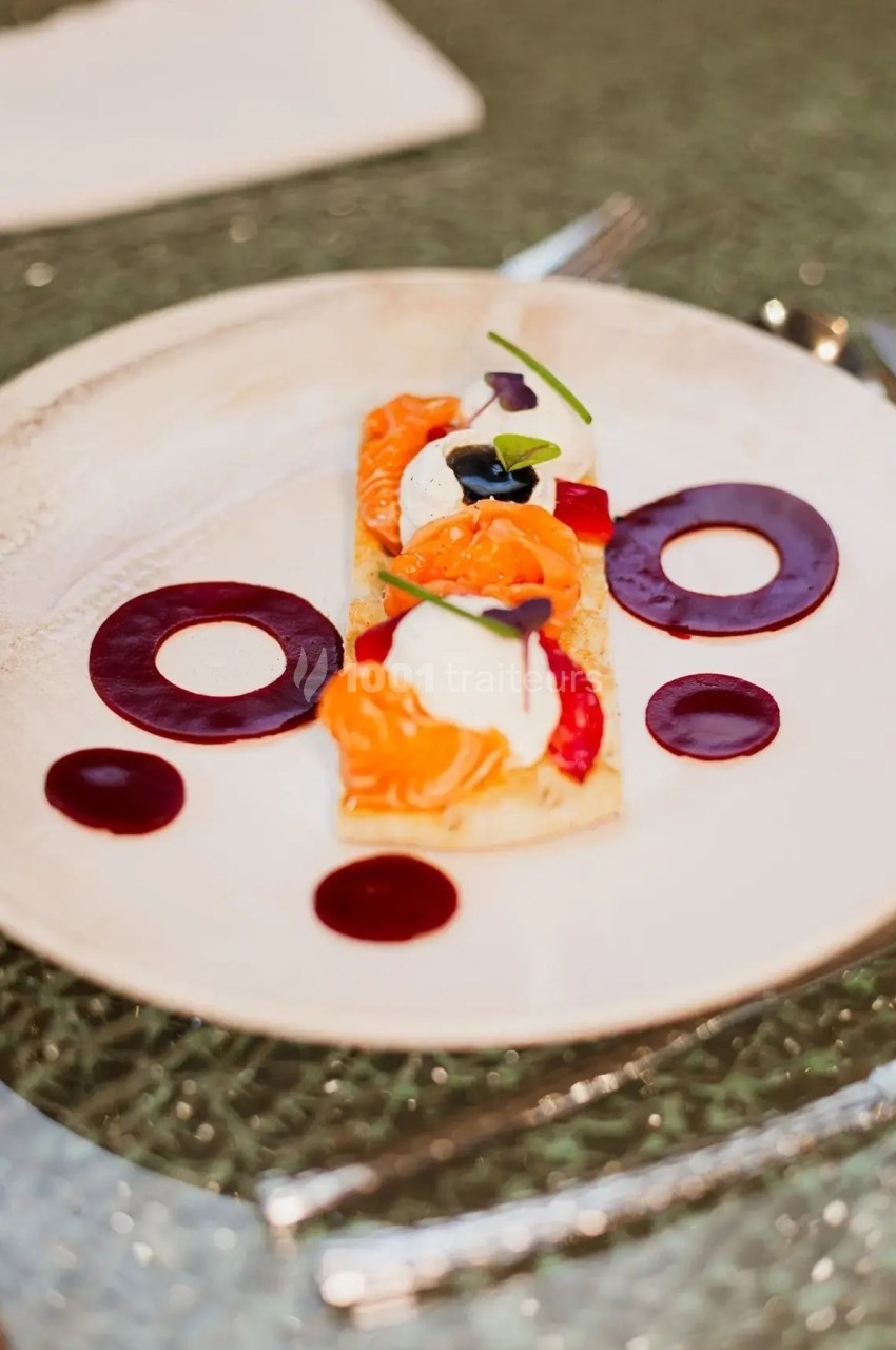 Assiette gastronomique avec saumon, crème, légumes colorés et cercles de sauce rouge, présentée sur une table élégante.