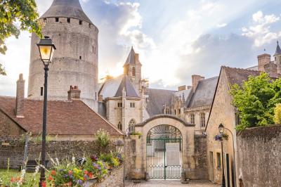 Un homme marche dans la cour d'un château médiéval avec une tour ronde et des bâtiments en pierre sous un ciel bleu.