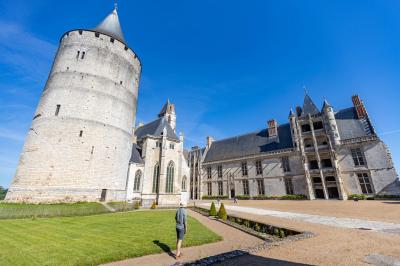Un homme marche dans la cour d'un château médiéval avec une tour ronde et des bâtiments en pierre sous un ciel bleu.