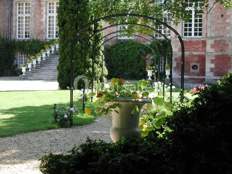 Jardin avec une allée bordée d'arches métalliques, un grand pot de fleurs au centre et un bâtiment en briques en arrière…