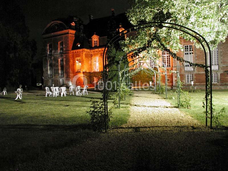 Allée éclairée de nuit avec des arches végétalisées, menant à un bâtiment ancien en briques illuminé.