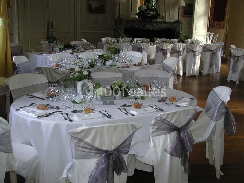 Tables rondes élégamment dressées avec nappes blanches, chaises décorées de rubans gris, et centres de table floraux.