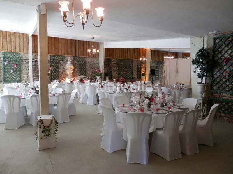 Salle de réception décorée avec des tables rondes, nappes blanches et chaises couvertes, ambiance lumineuse et fleurie.