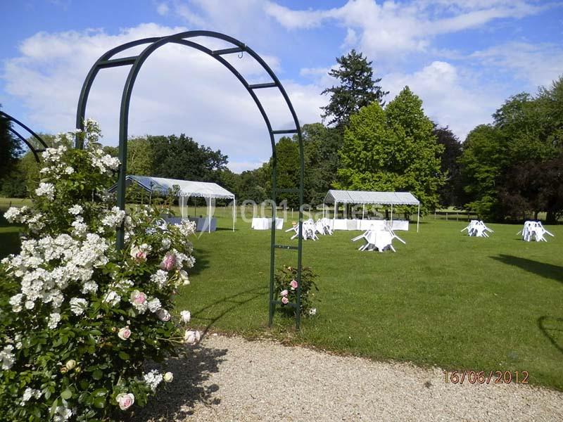 Vue d'un jardin avec des arches fleuries, des tables blanches et des tonnelles installées sur une pelouse.