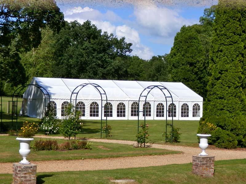 Tente blanche avec fenêtres en arc installée sur une pelouse entourée d'arbres et de parterres fleuris.