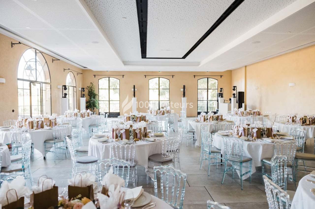 Salle de réception lumineuse avec tables rondes dressées, chaises transparentes et grandes fenêtres en arc.