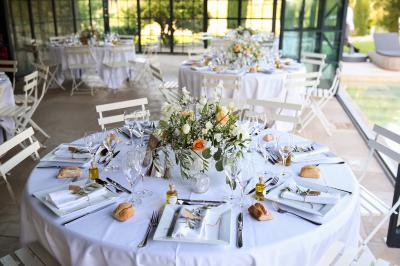 Table dressée avec nappes blanches, verres à vin, couverts, petits pains et centre de table floral dans une salle lumineuse.
