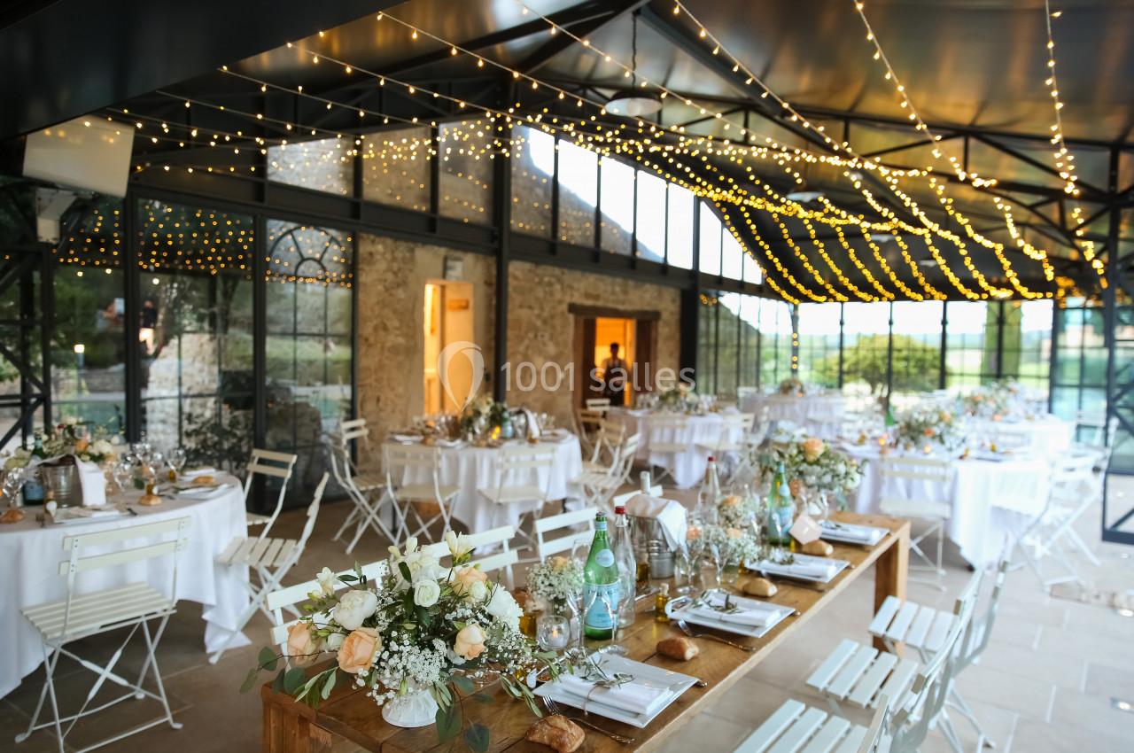 Salle de réception décorée avec des guirlandes lumineuses, des tables dressées et des chaises blanches.