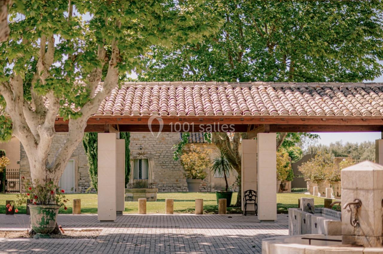Cour extérieure avec toit en tuiles, arbres ombrageants, pavés au sol et bâtiments en pierre en arrière-plan.