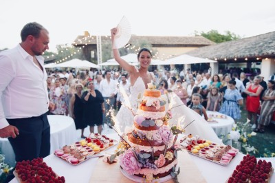 Une mariée souriante brandit un éventail devant un gâteau de mariage décoré, entourée d'invités en extérieur.