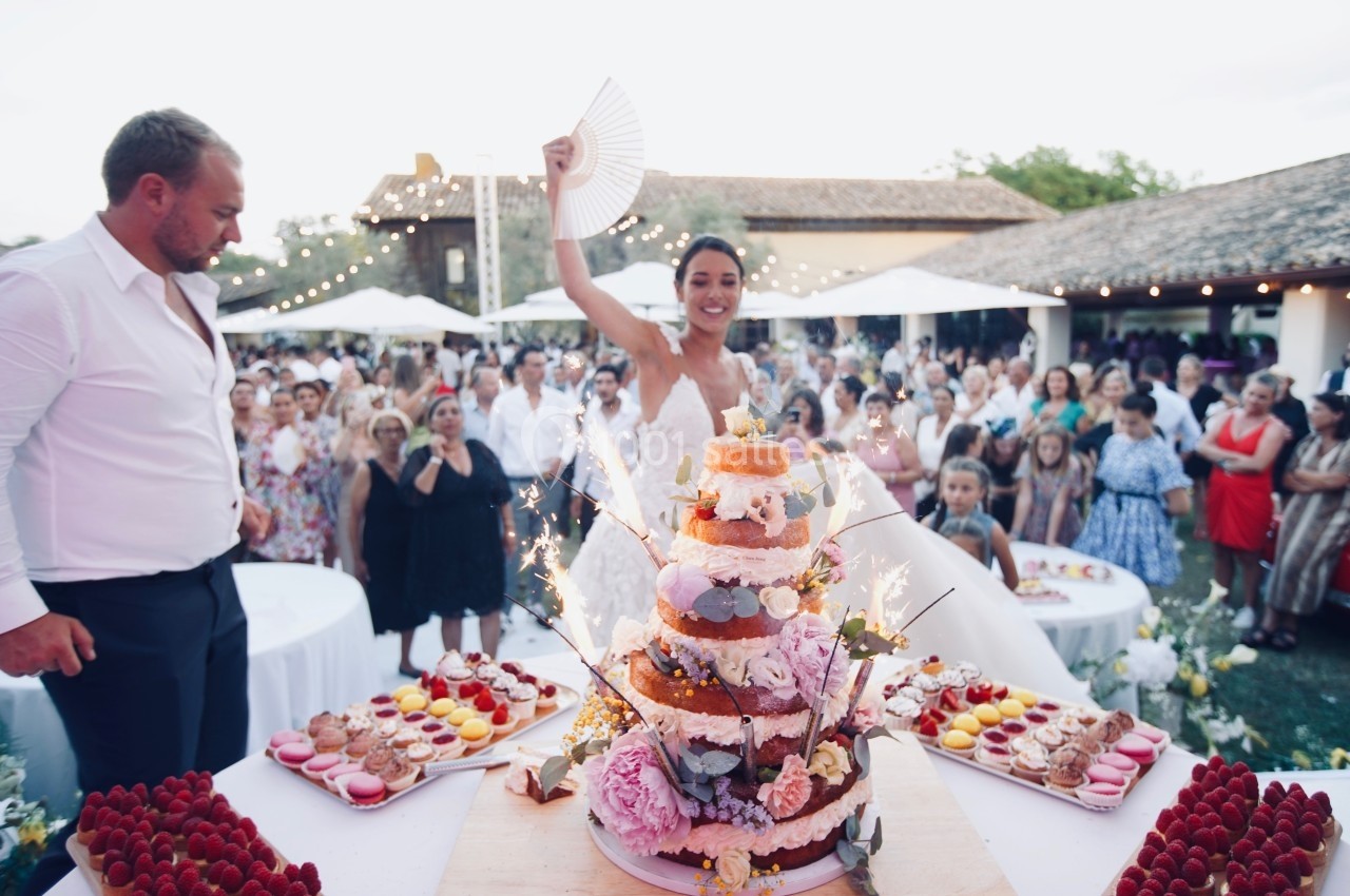 Une mariée souriante brandit un éventail devant un gâteau de mariage décoré, entourée d'invités en extérieur.