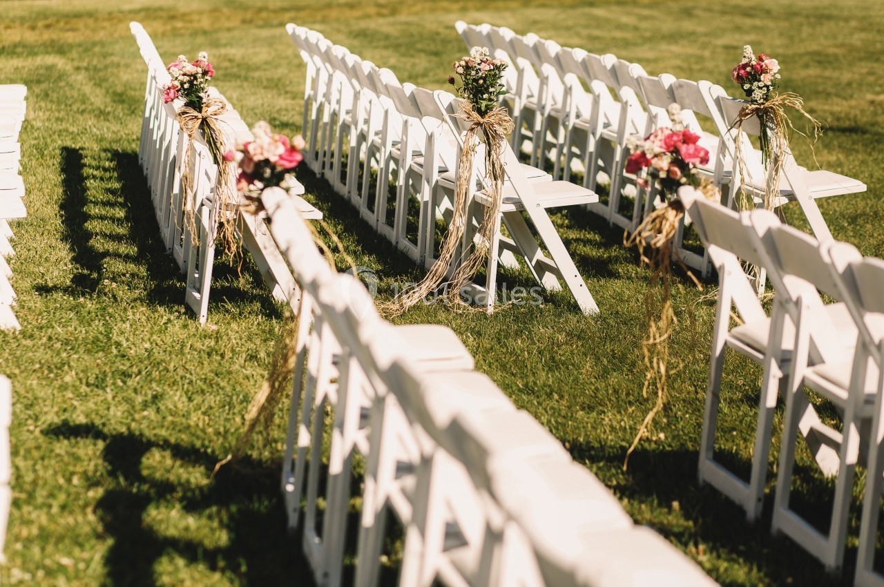 Chaises blanches alignées en extérieur, décorées de bouquets de fleurs roses et de rubans, sur une pelouse verte.