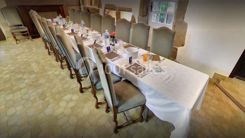 Table dressée avec nappes blanches, chaises alignées, vaisselle et verres, dans une salle au sol carrelé et murs en pierre.