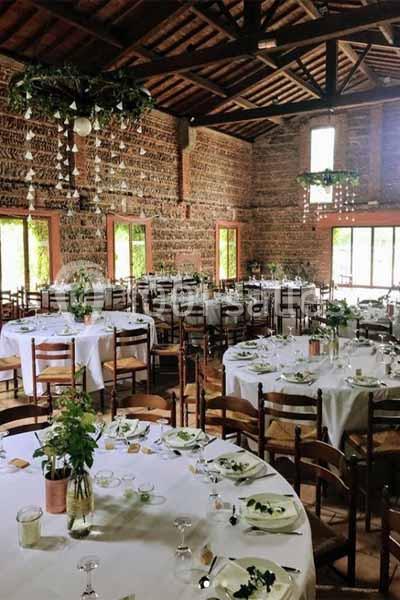 Salle de réception rustique avec tables rondes dressées, nappes blanches et décorations végétales suspendues.