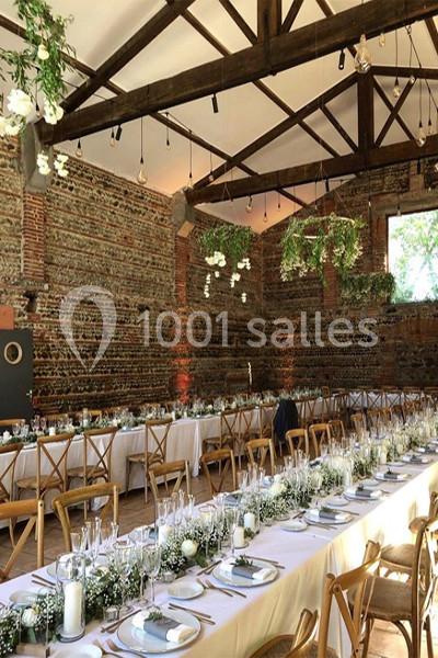 Grande salle de réception avec tables décorées de fleurs blanches et verdure, sous une charpente en bois apparente.
