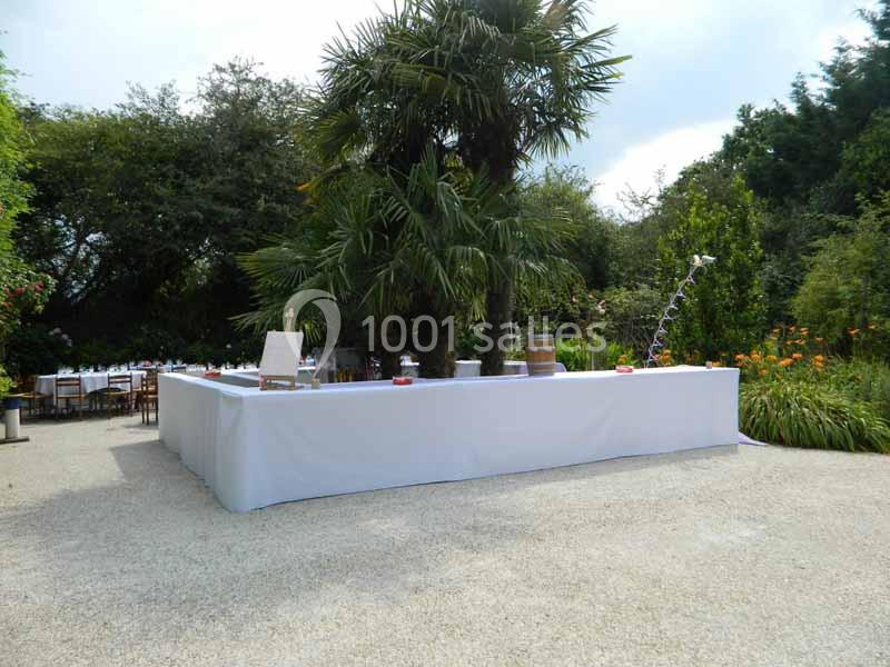 Bar extérieur couvert de nappes blanches, entouré de végétation luxuriante avec des tables en arrière-plan.