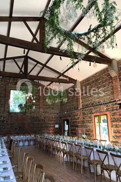 Grande salle de réception avec murs en briques, longues tables décorées et guirlandes végétales suspendues au plafond.