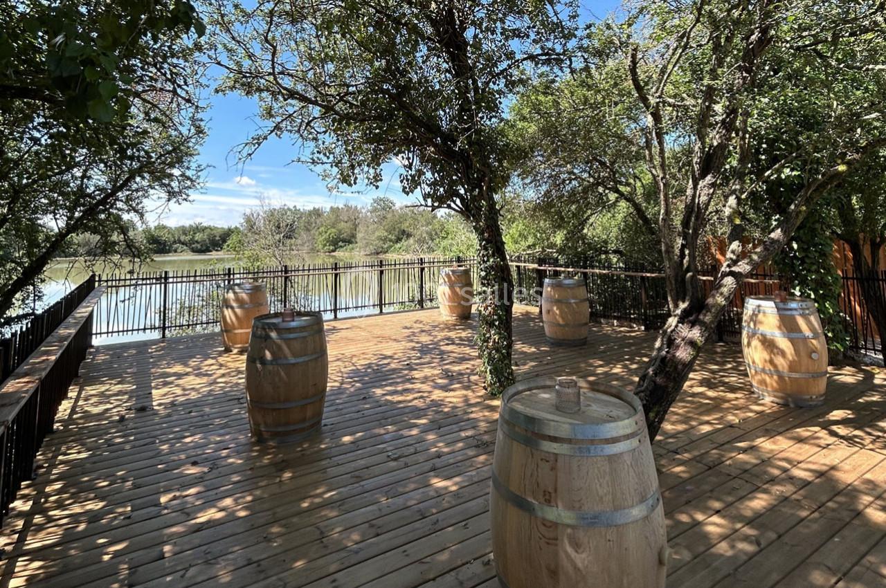 Terrasse en bois avec des tonneaux en décoration, entourée d'arbres et donnant sur un plan d'eau.