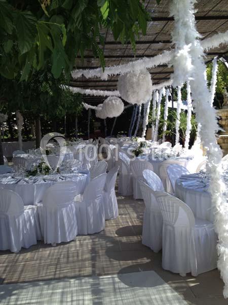 Salle extérieure décorée pour un événement, avec des tables rondes dressées, nappes blanches et ornements suspendus.