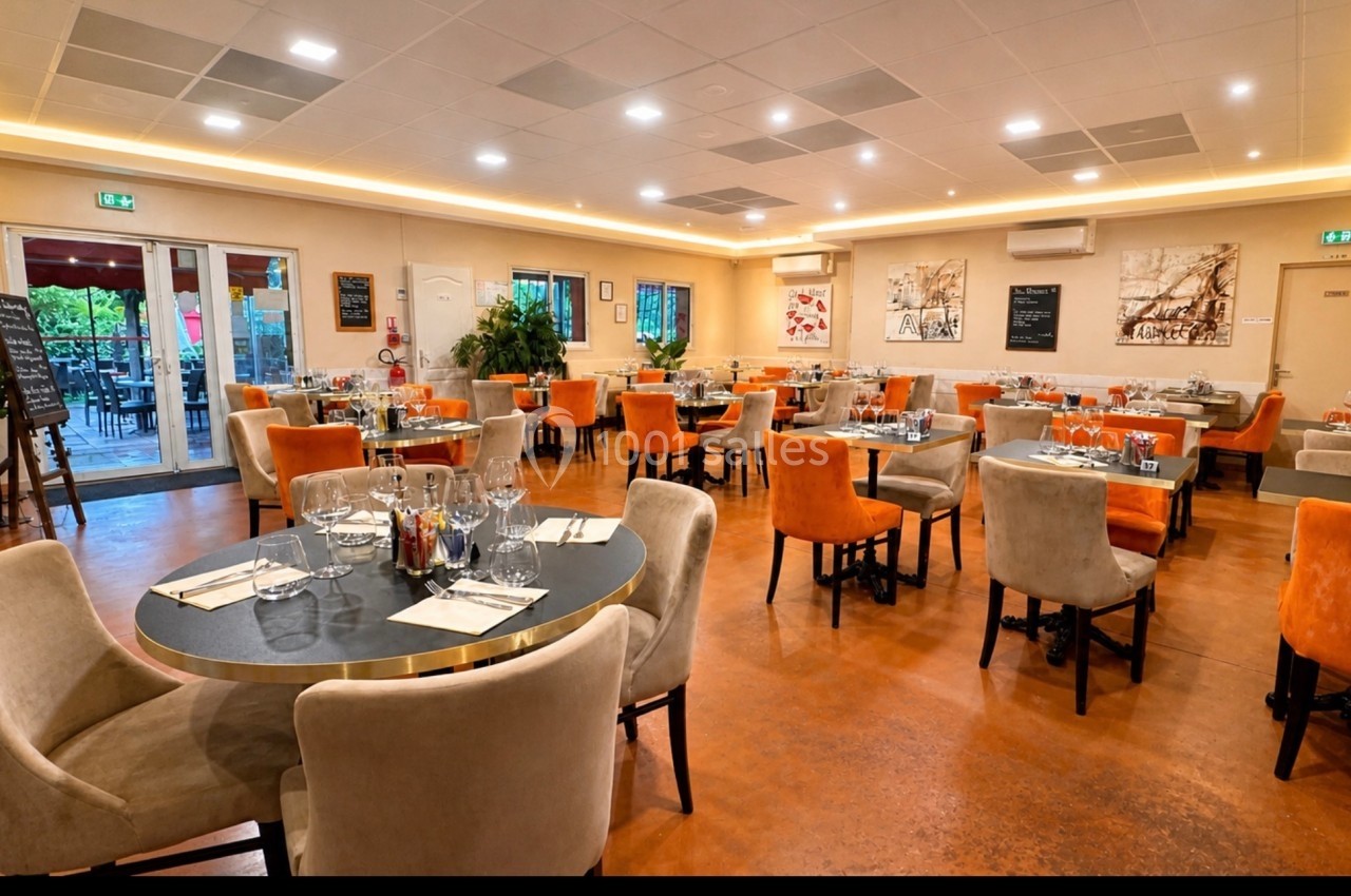 Salle de restaurant lumineuse avec tables dressées, chaises colorées et grandes baies vitrées donnant sur une terrasse.