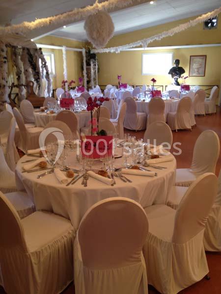 Salle de réception décorée avec des tables rondes, nappes blanches, chaises houssées et centres de table fleuris.