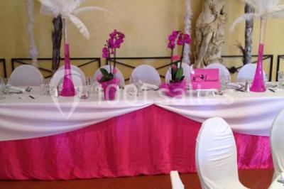 Table décorée avec une nappe blanche et rose, ornée de fleurs violettes et de plumes blanches, dans une salle festive.