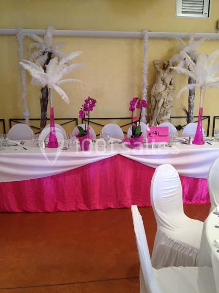 Table décorée avec une nappe blanche et rose, ornée de fleurs violettes et de plumes blanches, dans une salle festive.