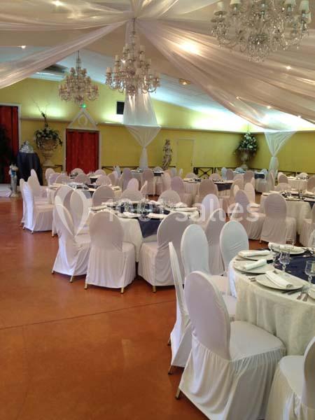 Salle de réception décorée avec des tables rondes dressées, nappes blanches et chaises houssées, sous un plafond drapé.