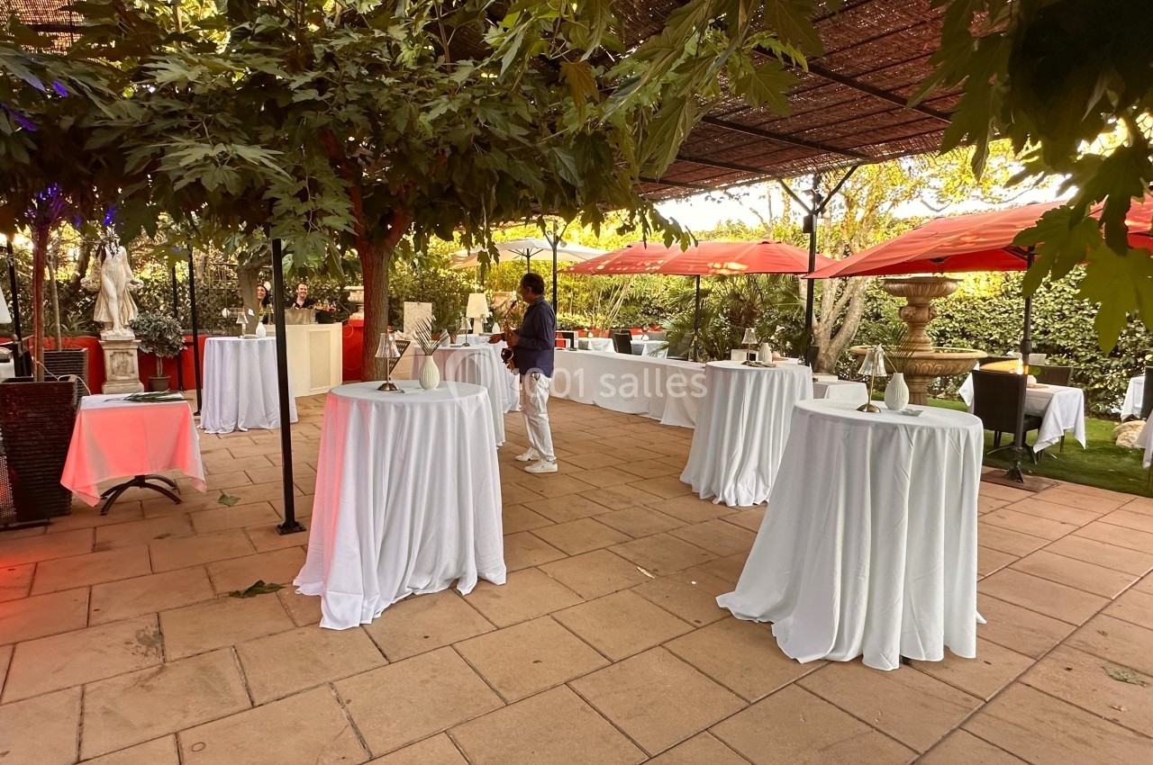 Terrasse aménagée avec des tables hautes drapées de blanc, des parasols rouges et une végétation abondante.