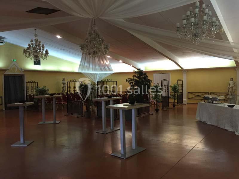 Salle de réception spacieuse avec tables hautes, chaises alignées, plantes décoratives et lustres suspendus.