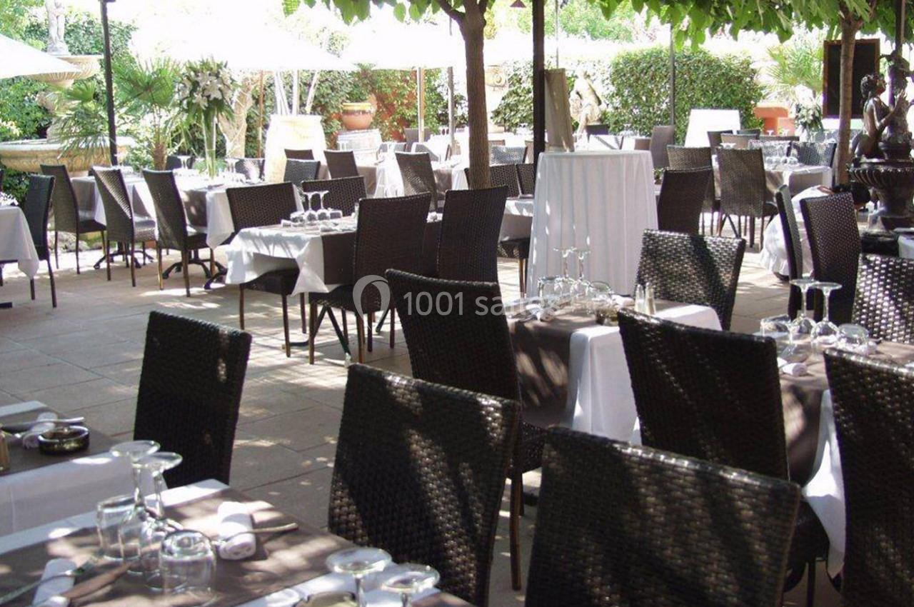 Terrasse d'un restaurant avec des tables dressées, chaises en osier, nappes blanches et végétation en arrière-plan.