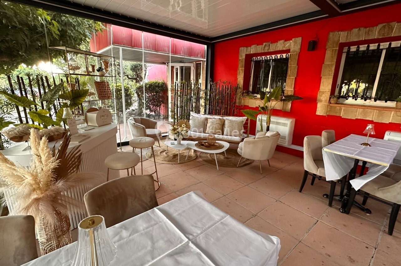 Terrasse couverte avec tables dressées, fauteuils, plantes décoratives et mur rouge en arrière-plan.