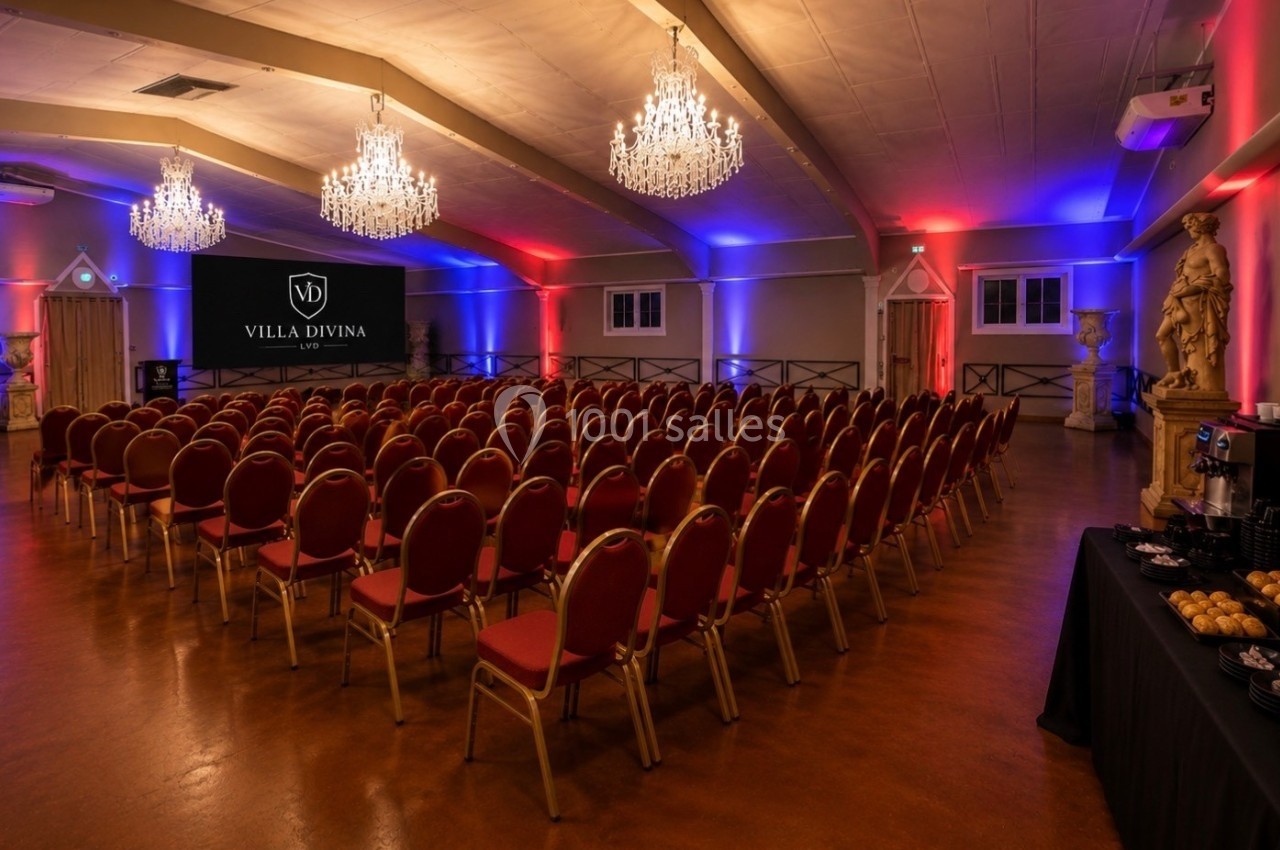 Salle de réception avec rangées de chaises rouges, éclairage coloré et écran affichant ’Villa Divina’.