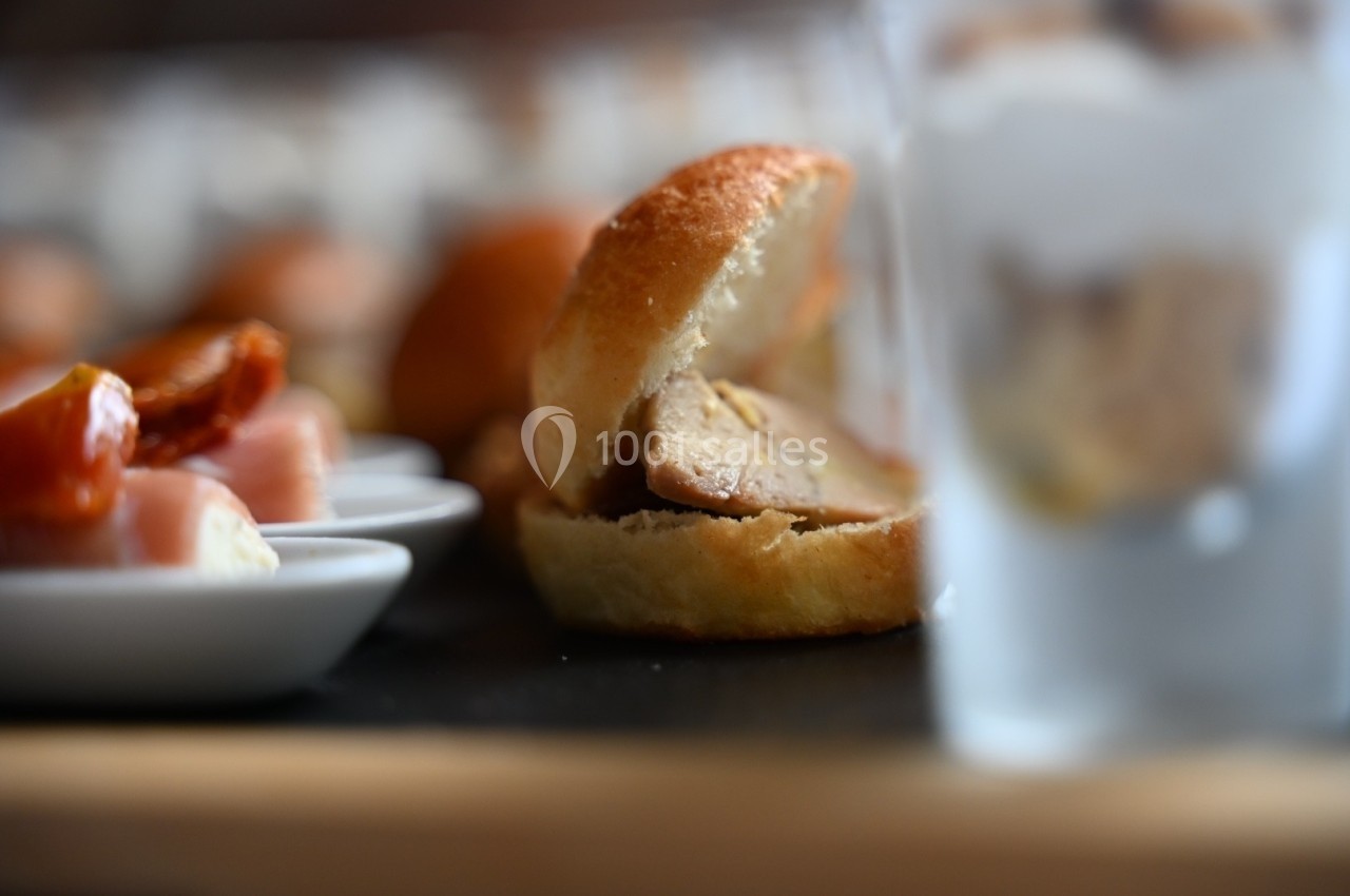 Mini sandwich au foie gras sur une planche, accompagné de tomates séchées et de verrines floues en arrière-plan.
