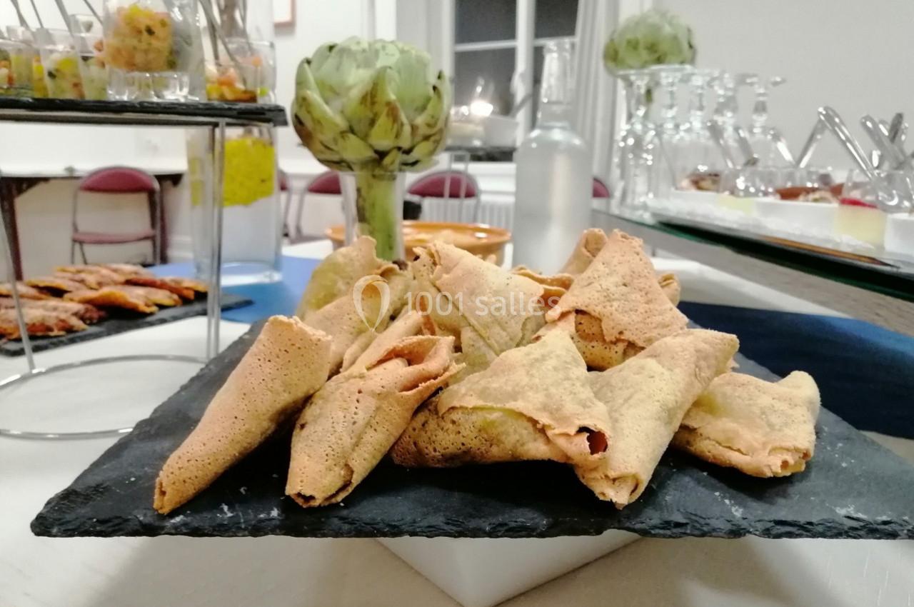 Assortiment de samoussas disposés sur une ardoise, entourés de verrines et d'un artichaut en décoration.